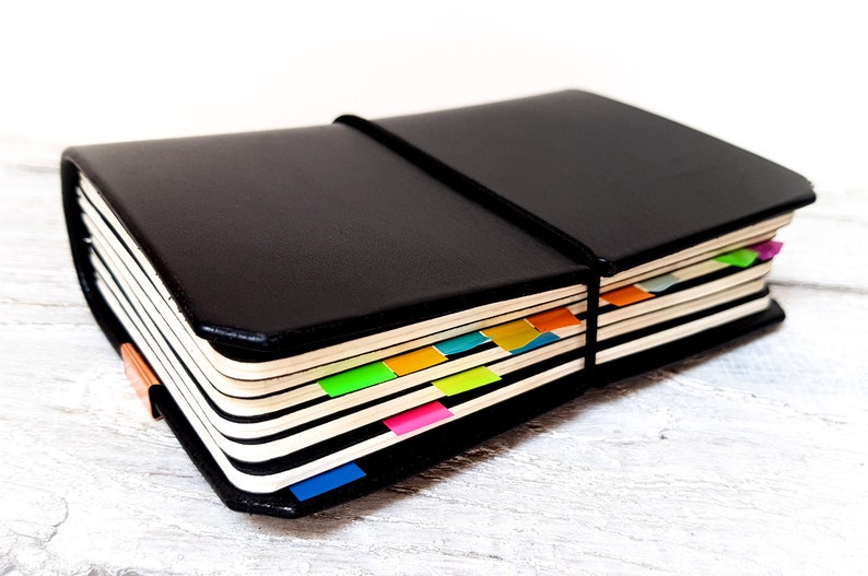 May include: A black leather notebook with a black elastic band. The pages are marked with colourful sticky tabs in orange, green, pink, and blue. The notebook is on a white wooden surface, with a light wood grain.