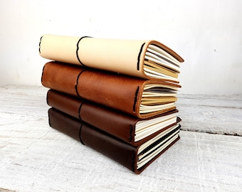 May include: A stack of four leather-bound journals in different shades of brown, beige, and black. The journals are closed and have a simple, elegant design.