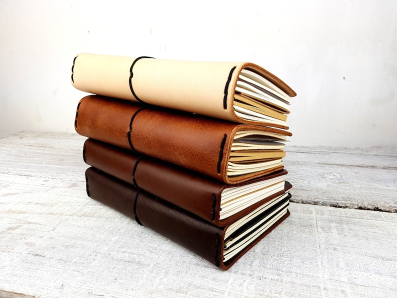 May include: A stack of four leather-bound journals in different shades of brown, beige, and black. The journals are closed and have a simple, elegant design.