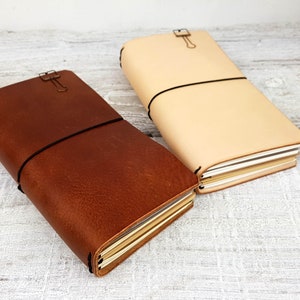 May include: Two leather-bound journals, one brown and one beige, with elastic closures and metal clasps. The journals are closed and stacked on top of each other.