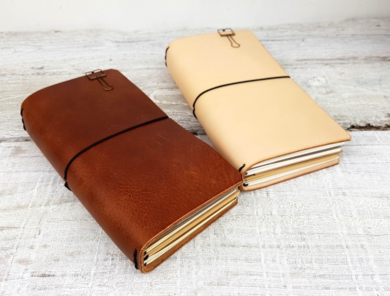 May include: Two leather-bound journals, one brown and one beige, with elastic closures and metal clasps. The journals are closed and stacked on top of each other.