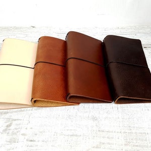May include: Four leather journals in varying shades of cream, tan, brown, and dark brown. Each journal is closed with a black elastic band. The journals are arranged side by side, showcasing the different color options.