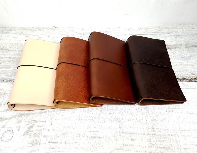 May include: Four leather journals in varying shades of cream, tan, brown, and dark brown. Each journal is closed with a black elastic band. The journals are arranged side by side, showcasing the different color options.