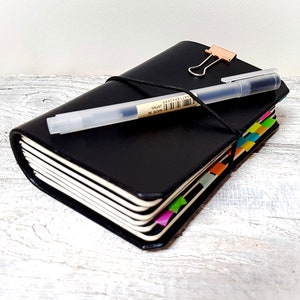 May include: A black leather journal with a pen and a rose gold binder clip. The journal has colourful page markers and a black elastic closure. The pen is clear with black ink and has text on it.
