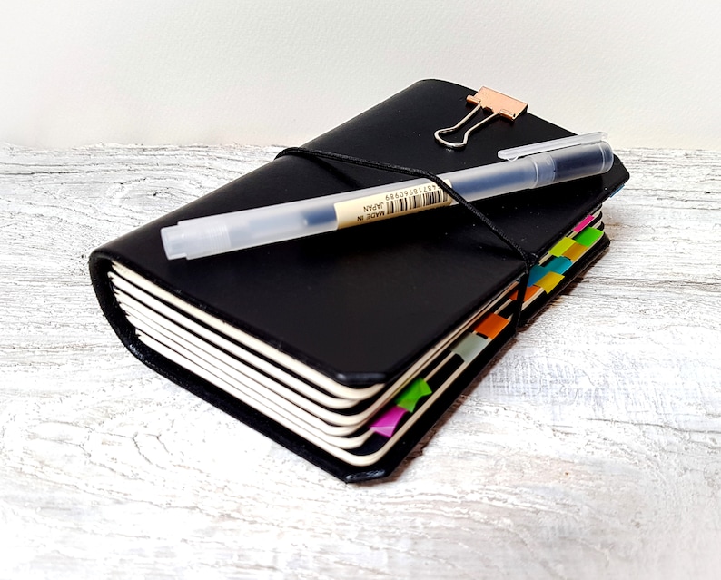 May include: A black leather journal with a pen and a rose gold binder clip. The journal has colourful page markers and a black elastic closure. The pen is clear with black ink and has text on it.