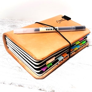 May include: A tan leather journal with a black elastic closure and a black pen on top. The journal has colorful page dividers and a black clip. The pen has the text "MUJI" printed on it.