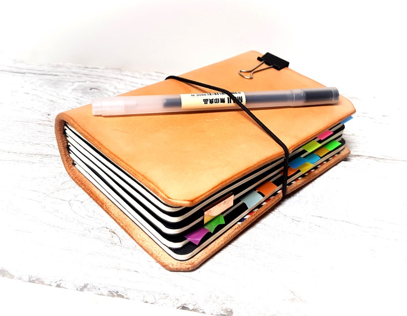 May include: A tan leather journal with a black elastic closure and a black pen on top. The journal has colorful page dividers and a black clip. The pen has the text "MUJI" printed on it.