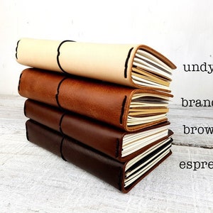 May include: Four leather journals in varying shades: undyed, brandy, brown, and espresso. Each journal features cream-coloured pages and black stitching. The image includes text on the right, indicating the colour of each journal.