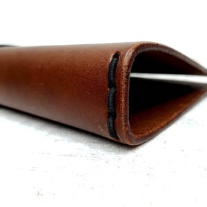 May include: A brown leather notebook cover with black stitching. The cover is folded in half and the stitching is visible on the edge.