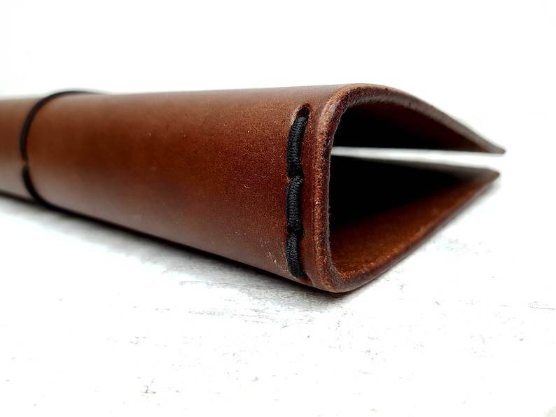 May include: A brown leather notebook cover with black stitching. The cover is folded in half and the stitching is visible on the edge.