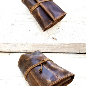 Puede incluir: Una bolsa de cuero marr&oacute;n para pipa con cierre de corbata. La bolsa est&aacute; doblada y atada con una correa de cuero. La bolsa est&aacute; sobre una superficie de madera blanca. "Pipe Pouch fully loaded" est&aacute; escrito en texto negro sobre la bolsa.