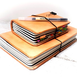 May include: Two stacked, tan leather journals with black and white striped pages. A black elastic band secures the top journal, with a pen and a binder clip on top. Colourful page markers are visible.