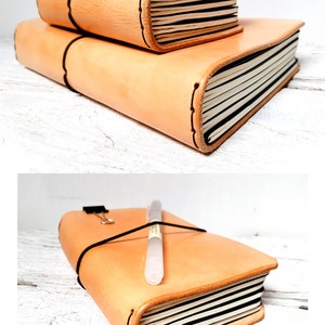 May include: Two stacked, light brown leather journals with black elastic closures. The top journal is smaller than the bottom one. The pages are white with black lines. A pen and a binder clip rest on the larger journal.