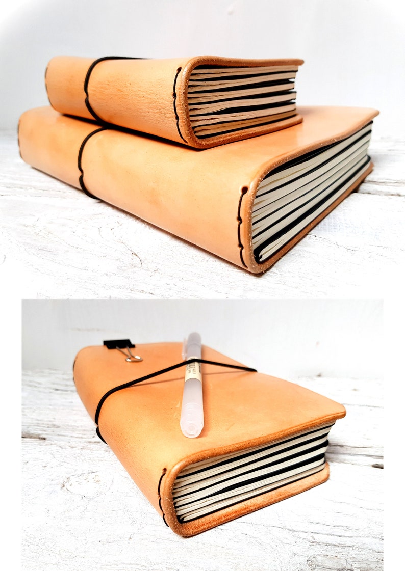 May include: Two stacked, light brown leather journals with black elastic closures. The top journal is smaller than the bottom one. The pages are white with black lines. A pen and a binder clip rest on the larger journal.