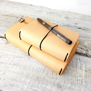 May include: Two beige leather journals with black stitching are stacked on top of each other. A grey pen is lying on top of the journals and is secured with a black elastic band.