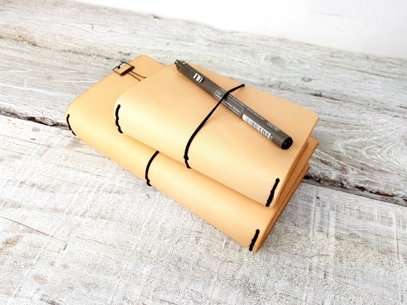 May include: Two beige leather journals with black stitching are stacked on top of each other. A grey pen is lying on top of the journals and is secured with a black elastic band.