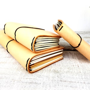 May include: Three leather-bound journals in various sizes, stacked on a white wooden surface. The journals are a light beige colour with black stitching and a decorative edge.