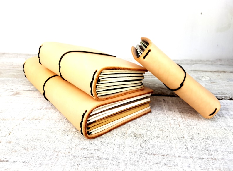 May include: Three leather-bound journals in various sizes, stacked on a white wooden surface. The journals are a light beige colour with black stitching and a decorative edge.