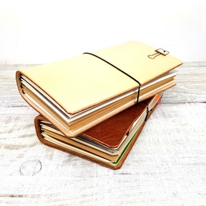 May include: Two leather-bound journals stacked on top of each other. The top journal is cream-coloured and has a black elastic band around it. The bottom journal is brown and has a green ribbon bookmark.