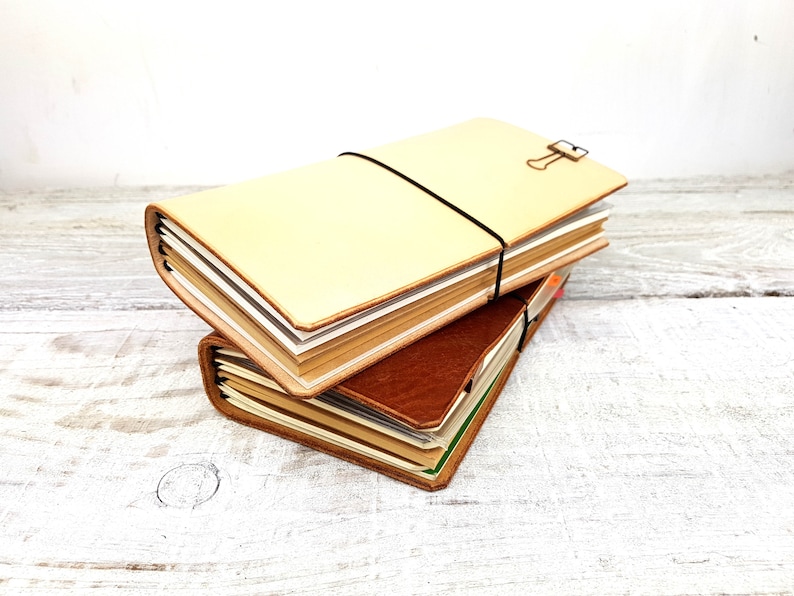 May include: Two leather-bound journals stacked on top of each other. The top journal is cream-coloured and has a black elastic band around it. The bottom journal is brown and has a green ribbon bookmark.