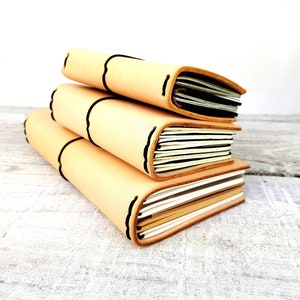 May include: Three leather-bound journals stacked on a white wooden surface. The journals are beige with black stitching and have a natural, rustic look.