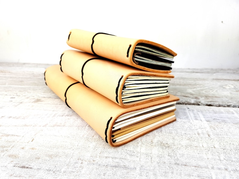 May include: Three leather-bound journals stacked on a white wooden surface. The journals are beige with black stitching and have a natural, rustic look.