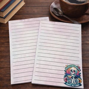 May include: Two lined notepads with a colourful design and the text "AT LEAST MY TRAUMA MADE ME FUNNY". A cup of coffee and books are in the background. The notepads are on a wooden surface.