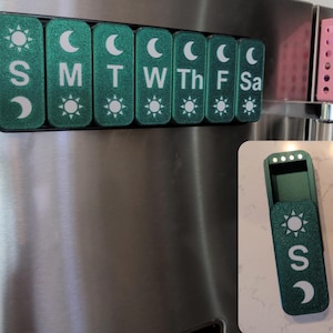 May include: A magnetic weekly pill organizer in shades of green, with each day labeled with a letter and a sun and moon design. The organizer is attached to a stainless steel refrigerator. Two additional open pill organizers are shown.