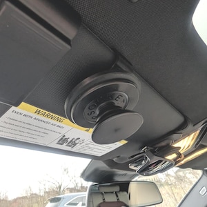 May include: A black PopSockets phone grip attached to a car visor. The grip is round with the letters "SO" and "E" visible. A warning label is partially visible.