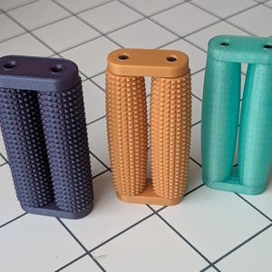 May include: Three colorful, textured, dual-grip handles in purple, orange, and teal. Each handle has a rectangular top with two holes and two rounded, textured columns. The handles are designed for a comfortable grip and are likely for a bicycle or exercise equipment.