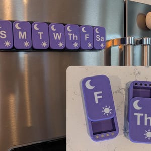 May include: A set of purple magnetic pill organizers labeled with days of the week, including S, M, T, W, Th, F, and Sa. Each unit features a white crescent moon and sun icon. Two individual units are shown open, revealing internal compartments.
