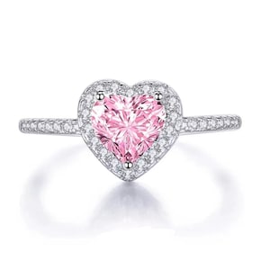 May include: A silver ring featuring a pink heart-shaped gemstone surrounded by a halo of smaller, clear stones. The band is also adorned with small, clear stones. The ring is set against a white background.