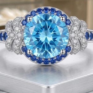 May include: A silver ring featuring a large, light blue gemstone at the centre, encircled by smaller blue stones. The band is adorned with more blue stones and white accents, creating a sparkling, elegant design.