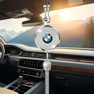 Op de afbeelding: Een zilveren auto-hanger met het BMW-logo in het midden, omringd door heldere kristallen. De hanger hangt aan een zwart koord en heeft een decoratieve kristallen kwast. De achtergrond toont een auto-interieur en een landschap.