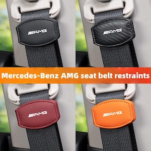 May include: Four Mercedes-Benz AMG seat belt restraints in black, carbon fibre, burgundy, and orange. Each restraint has the AMG logo in white. The restraints are attached to grey seat belts.