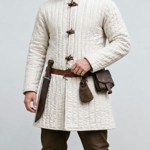 May include: A long-sleeved, off-white quilted tunic featuring a high collar and three brown toggle closures down the front. A brown leather belt is worn at the waist, holding a large knife in a sheath, a small brown pouch, and a rectangular leather bag.