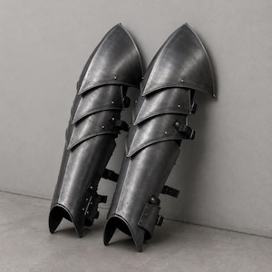 May include: A pair of dark grey metal greaves featuring a multi-layered articulated plate design. Each piece is constructed with overlapping steel segments secured by visible rivets and black leather straps with metal buckles for fastening.