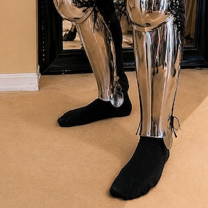 May include: Polished steel leg armor, featuring knee and shin protection. The armor is secured with leather straps and rivets. The lower legs are covered with black socks. The armor reflects the surrounding environment.