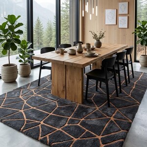 May include: A rectangular area rug featuring a dark grey, textured pile with a geometric pattern of intersecting copper-coloured lines. The rug is positioned on a concrete floor beneath a wooden dining table and black chairs in a room with large windows.