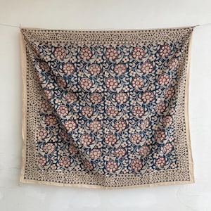 May include: A square textile featuring a repeating floral pattern in dark blue, cream, and muted red. The central design consists of stylized blossoms and leaves, framed by a decorative border with a geometric pattern in dark brown and cream.