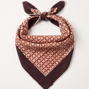 May include: A square bandana tied in a knot, featuring a deep plum border and a central panel with a repeating pattern of cream-coloured floral shapes and cross symbols on a terracotta background, finished with a light-coloured hemmed edge.
