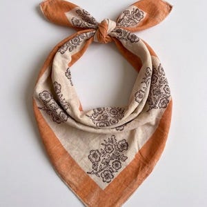 May include: A square bandana with a floral design. The bandana has a cream-coloured centre with a brown floral pattern and a burnt orange border. It is tied in a knot at the top.
