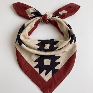 May include: A square bandana with a geometric design. The bandana features a cream-colored center with black and red geometric shapes and a red border. The bandana is tied at the top, forming a knot and two pointed ends.