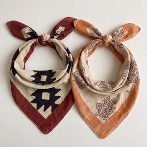 May include: Two triangular fabric bandanas tied in knots at the top. The left bandana features a cream base with a dark brown geometric pattern and a deep red border. The right bandana has a cream base with a brown floral print and orange borders.