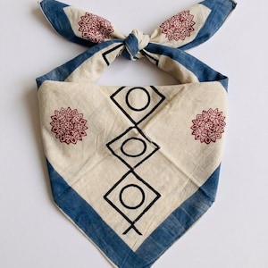 May include: Cream-colored bandana with a blue border and accents. The bandana features a repeating pattern of black geometric shapes and maroon floral designs. The bandana is tied at the top, showcasing the blue and maroon patterns.