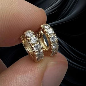May include: A pair of gold hoop earrings with multiple clear gemstones set along the front. The earrings are held between fingers against a dark, abstract background.
