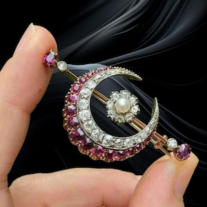 Pode incluir: Broche dourado em forma de meia-lua, com uma pérola central e diamantes. A curva da lua é adornada com pedras vermelhas. Pedras vermelhas e diamantes adicionais acentuam as extremidades do broche.