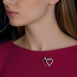 May include: A brooch featuring two interlocking heart-shaped frames. One heart is set with small, vibrant red stones, while the other heart is set with clear, sparkling white stones. The piece is pinned to a solid, deep magenta-colored fabric.