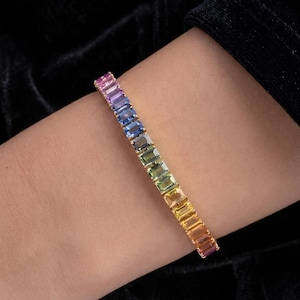 May include: A gold tennis bracelet featuring a row of rectangular baguette-cut gemstones arranged in a vibrant rainbow gradient. The stones transition through shades of purple, blue, green, yellow, orange, and red, set in a polished gold metal band.