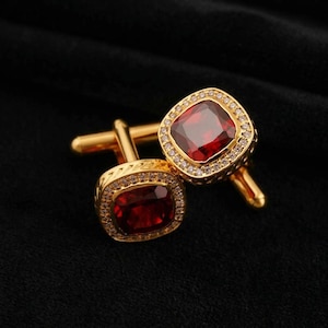 Puede incluir: Un par de gemelos dorados con una piedra central cuadrada de color rojo granate intenso. Cada gemelo está rodeado por un borde de pequeñas piedras claras y brillantes engastadas en un marco de metal dorado con un cierre de barra en T.